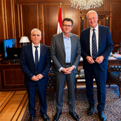 Three men in suits standing in a wood‑panelled office, smiling; a flag and chandelier in the background.