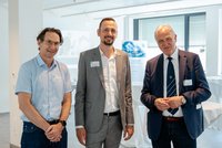 Drei Personen posieren bei einer Veranstaltung in Innenräumen; zwei tragen Anzug, eine Person Hemd und Jeans. Namensschilder sind sichtbar.