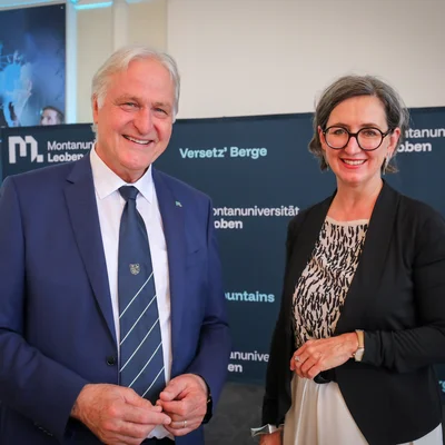 Zwei Personen in formeller Kleidung vor einem Montanuniversität‑Leoben‑Backdrop; links blauer Anzug mit Krawatte, rechts schwarzer Blazer.