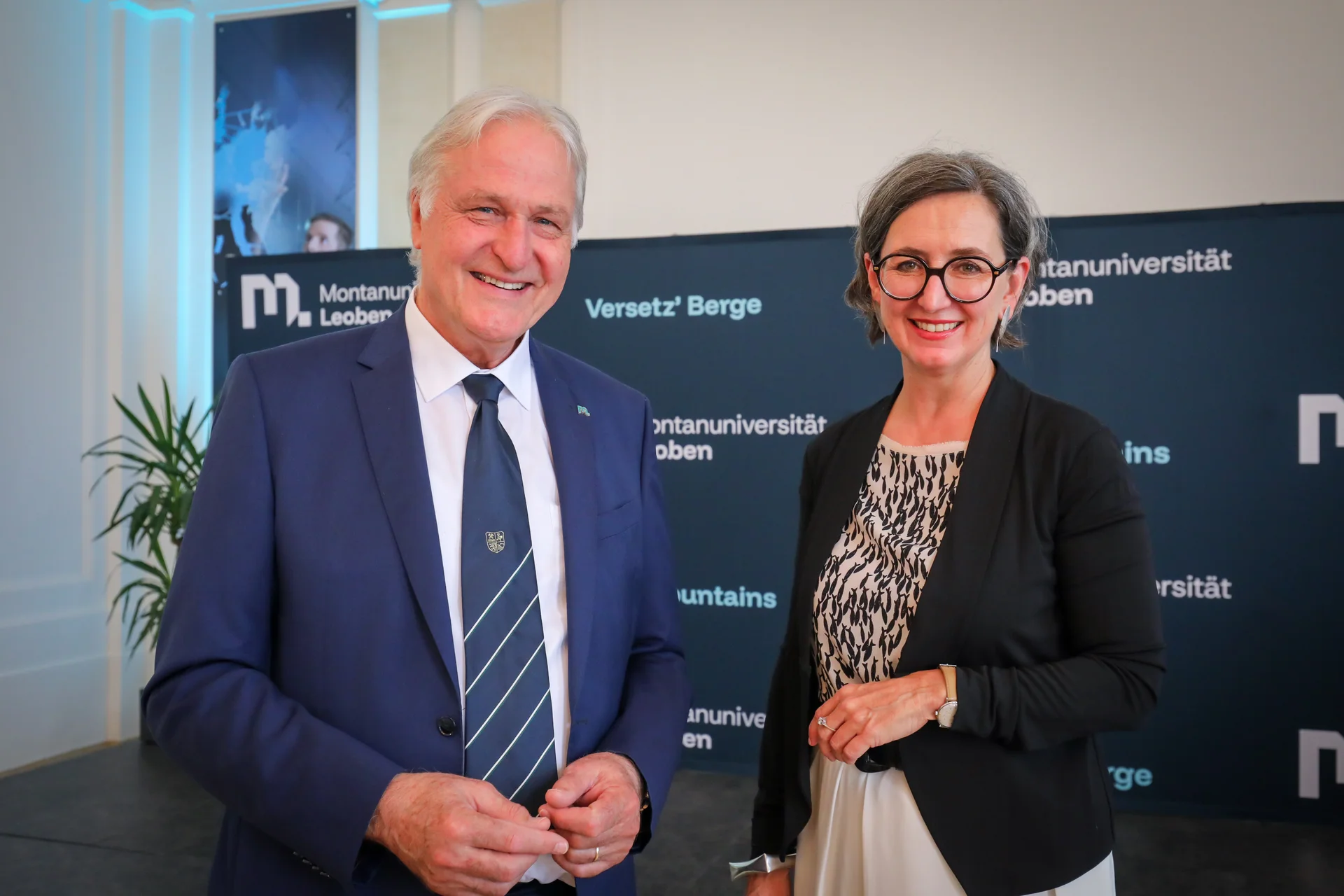 Zwei Personen in formeller Kleidung vor einem Montanuniversität‑Leoben‑Backdrop; links blauer Anzug mit Krawatte, rechts schwarzer Blazer.