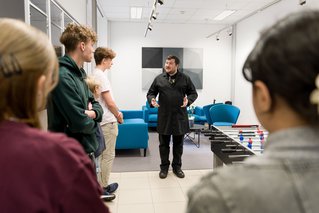 Group listening to a person in black clothing in a lounge area with sofas and a foosball table.