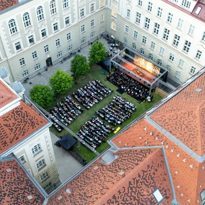 Zuschauerinnen sitzen bei einer Open‑Air‑Veranstaltung und blättern in Programmbroschüren; eine Person hält ein Getränk.