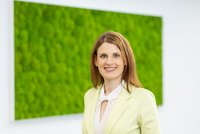      Porträt einer lächelnden Frau im hellgrünen Blazer vor einer großen grünen Mooswand in einem hellen Innenraum.