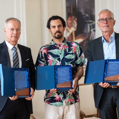 Drei Personen in einer Halle halten offene blaue Etuis mit Glas‑Awards. Zwei tragen Anzug, die mittlere Person ein buntes Hemd. Im Hintergrund Stuhlreihen.