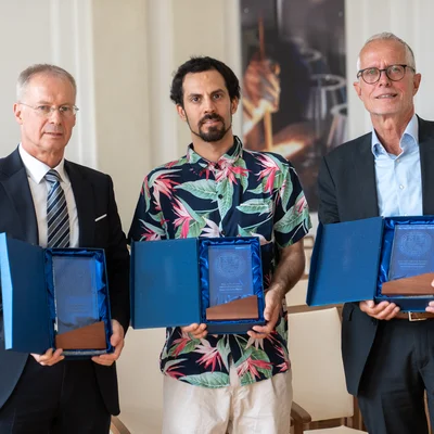 Drei Personen in einer Halle halten offene blaue Etuis mit Glas‑Awards. Zwei tragen Anzug, die mittlere Person ein buntes Hemd. Im Hintergrund Stuhlreihen.