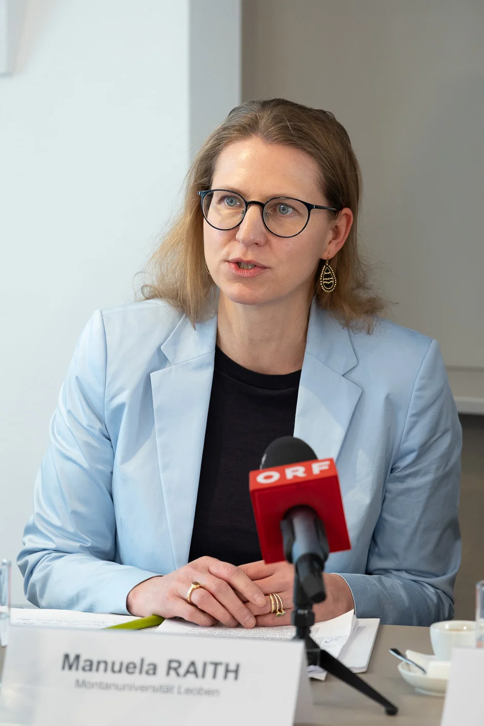 Eine Person sitzt bei einer Pressekonferenz oder einem Interview an einem Tisch. Sie trägt einen hellblauen Blazer über dunklem Oberteil, hat die Hände gefaltet und spricht vor einem roten Mikrofon mit ORF‑Logo. Vor ihr steht ein Namensschild; Unterlagen, Stifte und ein Glas sind auf dem Tisch.