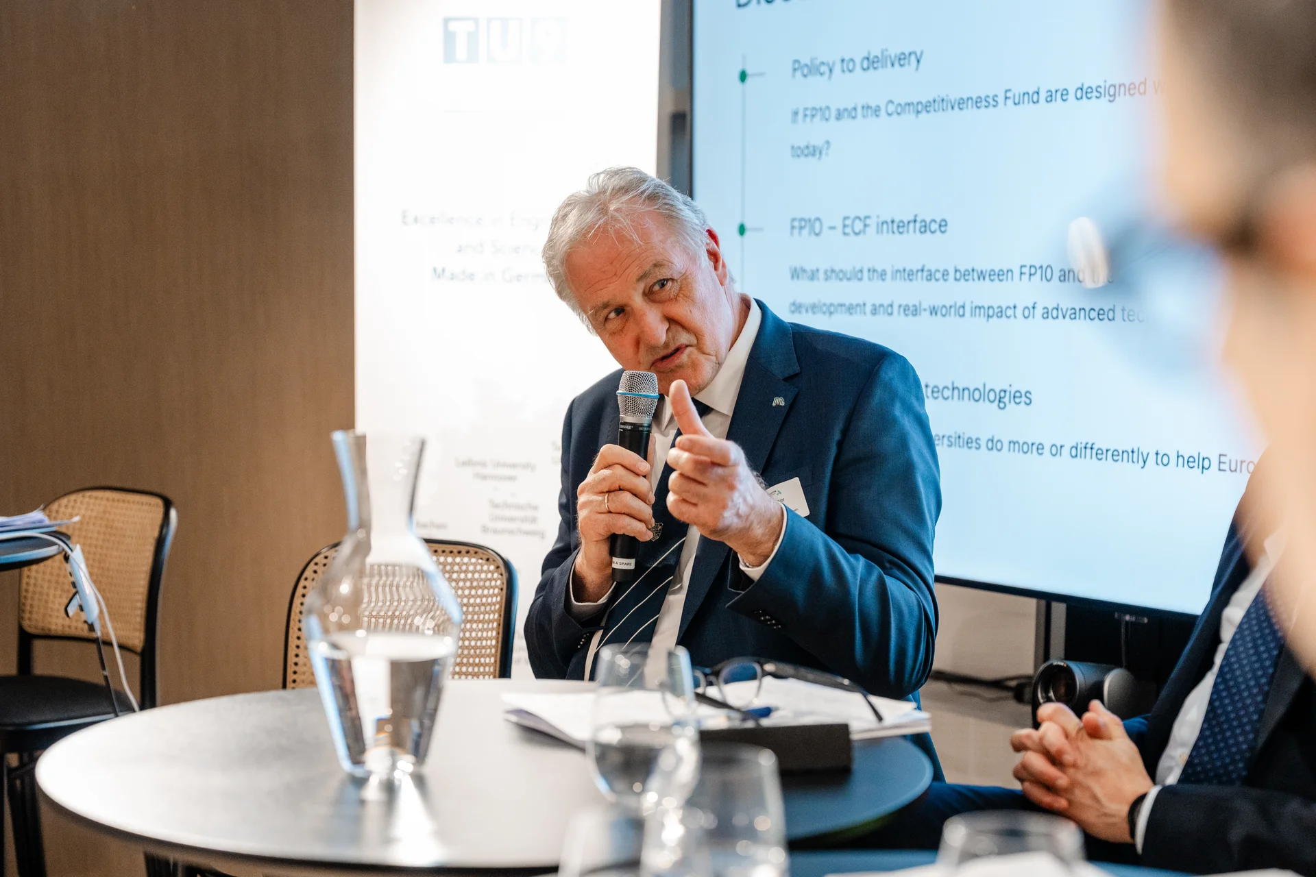 Rektor Peter Moser inmitten einer Podiumsdiskussion. Er hält ein Mikrofon in der Hand und zeigt einen Daumen nach oben.