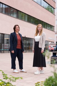 Zwei Personen in business‑casual Kleidung stehen auf einem gepflasterten Hof vor einem modernen, mehrgeschossigen Gebäude mit breiten Fensterbändern; Gesichter sind unkenntlich, im Vordergrund Pflanzen.