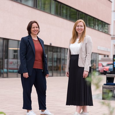 Zwei Personen in business‑casual Kleidung stehen auf einem gepflasterten Hof vor einem modernen, mehrgeschossigen Gebäude mit breiten Fensterbändern; Gesichter sind unkenntlich, im Vordergrund Pflanzen.