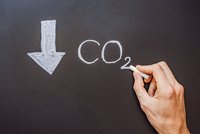 Hand schreibt mit Kreide „CO₂“ auf eine Tafel; daneben ein nach unten zeigender Pfeil – Symbol für sinkende CO₂-Emissionen.