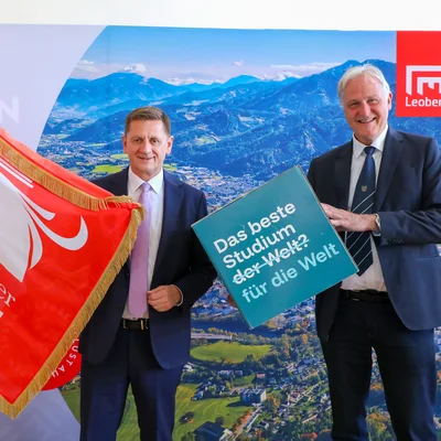 Zwei Männer im Anzug posieren vor einem Panorama‑Hintergrund mit Berg‑/Stadtansicht und Leoben‑Logo. Links wird eine rote Fahne „Österreichischer Städtebund“ gehalten. Rechts halten sie ein Schild mit der Aufschrift „Das beste Studium der Welt? für die Welt“. Gesichter sind unkenntlich gemacht.