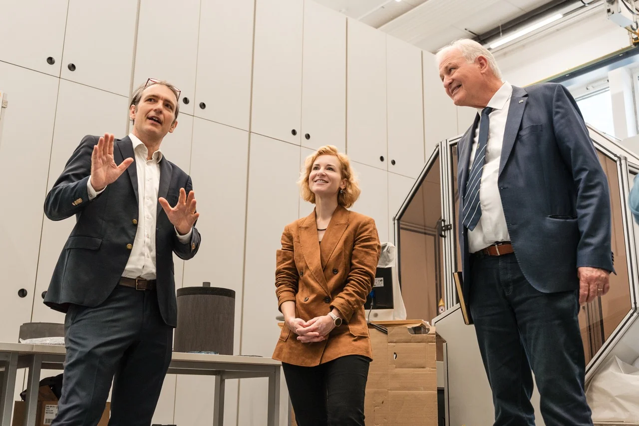 Drei Personen in Businesskleidung stehen in einem Labor/Technikraum; eine Person erklärt gestikulierend, die anderen hören zu. Im Hintergrund Schränke, Tische, Kartons und technische Geräte.