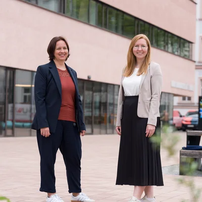 Zwei Personen in business‑casual Kleidung stehen auf einem gepflasterten Hof vor einem modernen, mehrgeschossigen Gebäude mit breiten Fensterbändern; Gesichter sind unkenntlich, im Vordergrund Pflanzen.