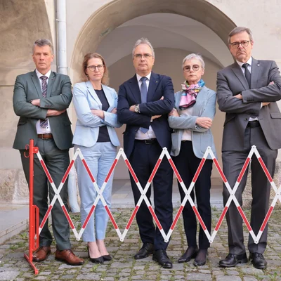 Fünf Personen in Businesskleidung stehen im Freien in einem historischen Hof mit Rundbögen. Alle haben die Arme verschränkt und posieren hinter einer ausgezogenen, rot‑weißen Scherensperre; Pflastersteine am Boden.
