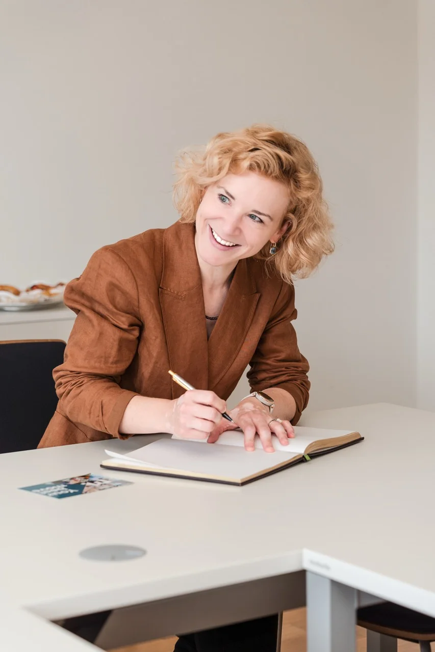 Person mit blondem Lockenhaar sitzt an einem Tisch und schreibt mit einem Stift in ein aufgeschlagenes Buch/ Gästebuch; sachliche Innenraumszene, helles Mobiliar.