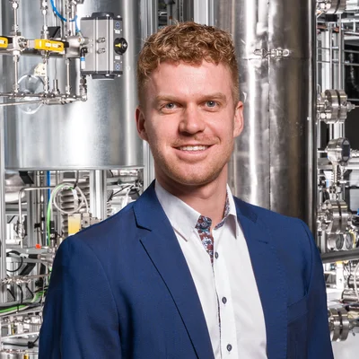 Person in blauem Anzug und weißem Hemd vor einer technischen Anlage mit Edelstahlrohren, Ventilen und Messinstrumenten – industrielles/pilotanlagenartiges Umfeld.
