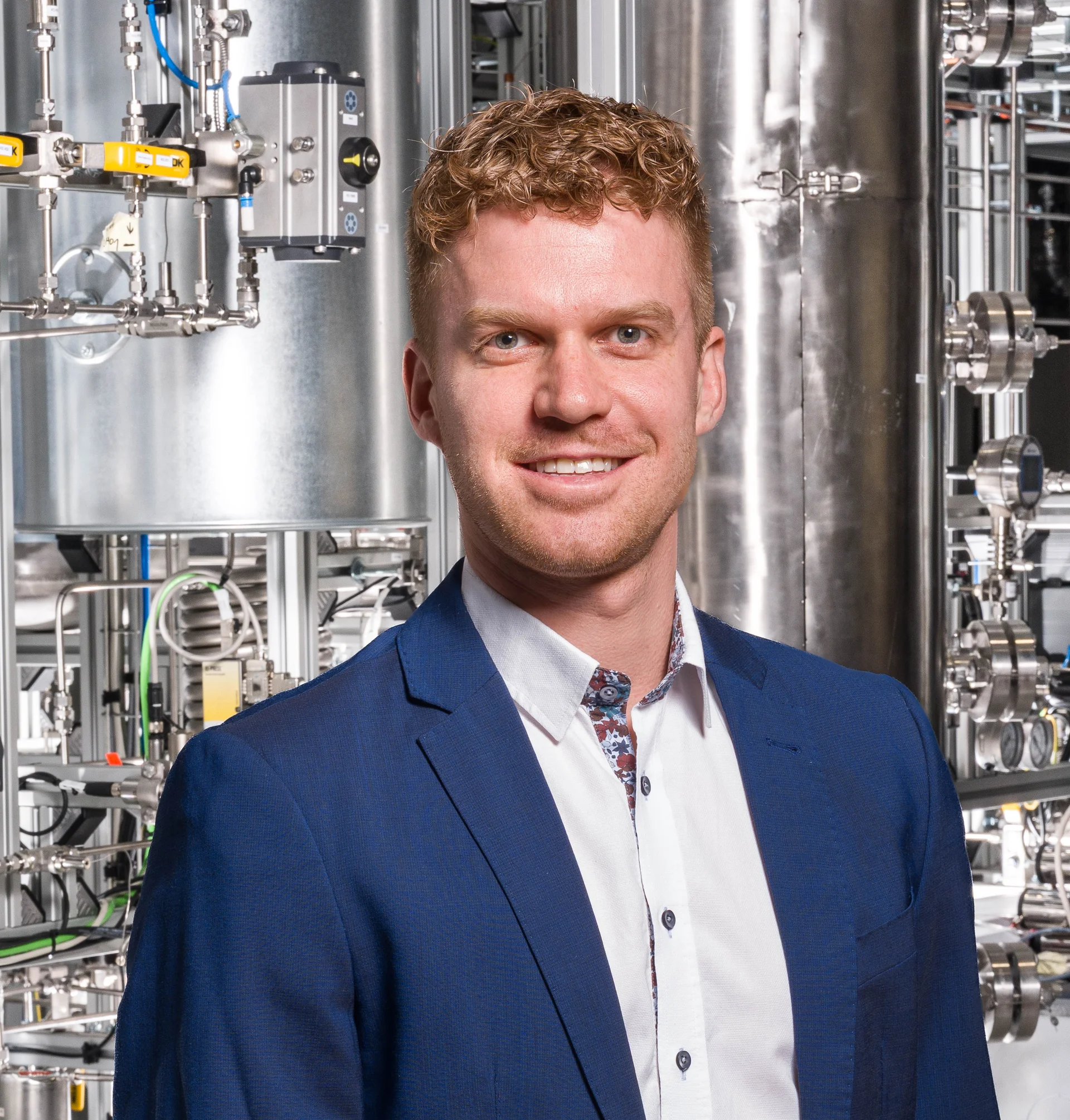 Person in blauem Anzug und weißem Hemd vor einer technischen Anlage mit Edelstahlrohren, Ventilen und Messinstrumenten – industrielles/pilotanlagenartiges Umfeld.