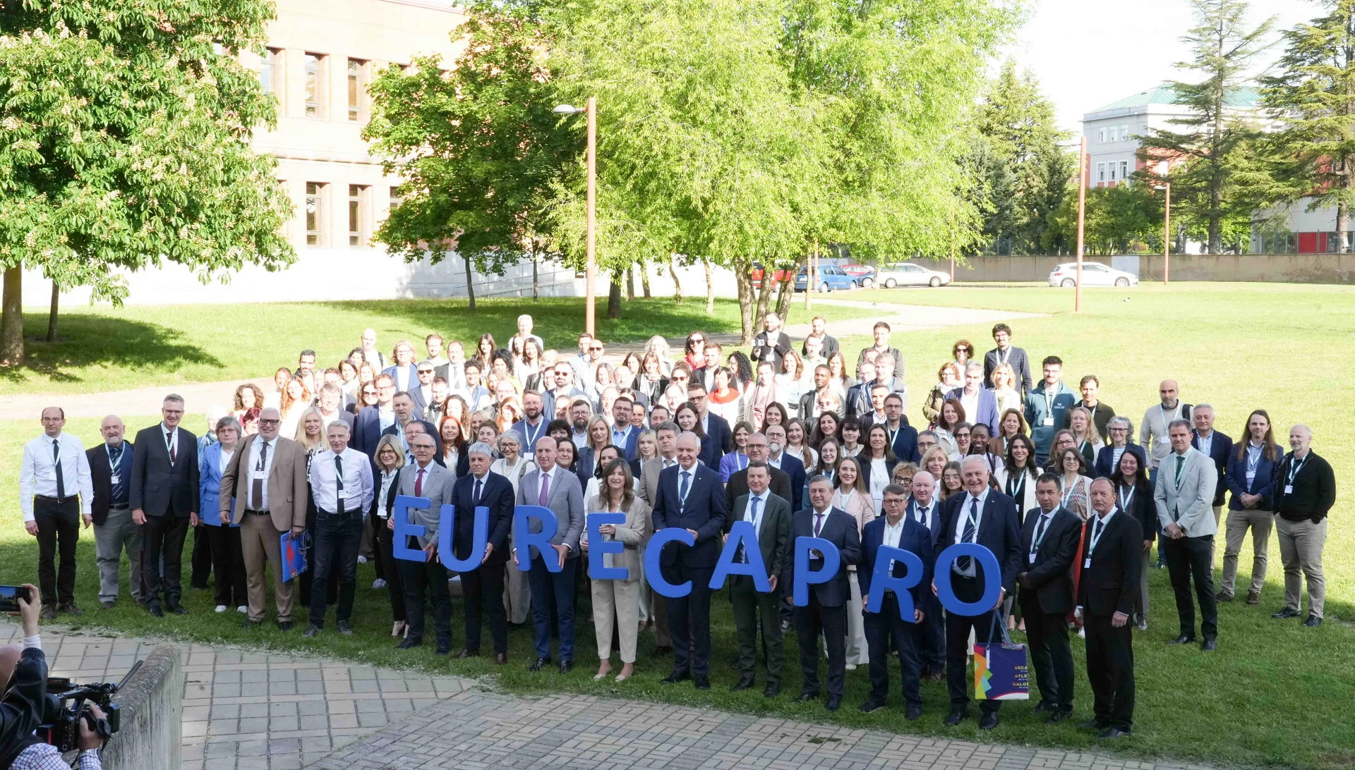 Großes Gruppenfoto im Freien auf einem Campusrasen. In der ersten Reihe halten Teilnehmende große blaue Buchstaben, die „EURECA PRO“ ergeben. Bäume und Hochschulgebäude im Hintergrund, sonniges Wetter.