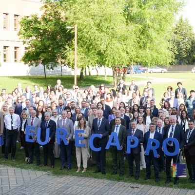 Großes Gruppenfoto im Freien auf einem Campusrasen. In der ersten Reihe halten Teilnehmende große blaue Buchstaben, die „EURECA PRO“ ergeben. Bäume und Hochschulgebäude im Hintergrund, sonniges Wetter.