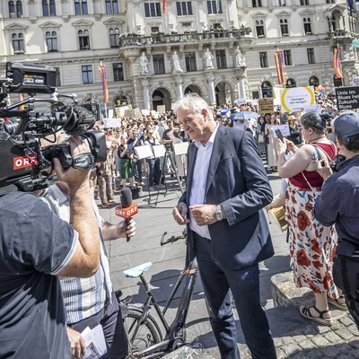 Presse‑ und Publikumsandrang auf einem Platz vor einem prächtigen historischen Rathaus. Kamerateams und Fotografen filmen, Menschen mit Transparenten und Fahnen stehen im Sonnenschein; vorne eine Person im Anzug, im Hintergrund die reich verzierte Fassade mit Turm.