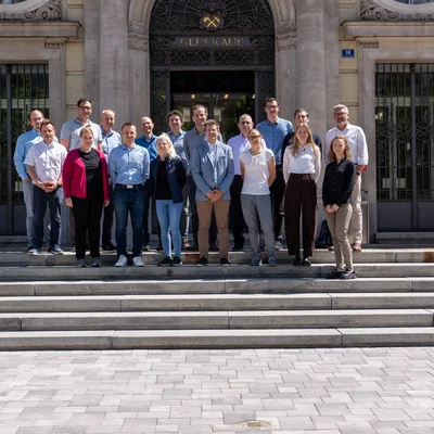 Gruppenfoto vor einem historischen Gebäude: Rund 18 Personen stehen auf breiten Steinstufen vor einer verzierten Eingangstür mit Gittertüren und sandfarbener Fassade. Sonniges Wetter, Schatten auf dem Pflaster.