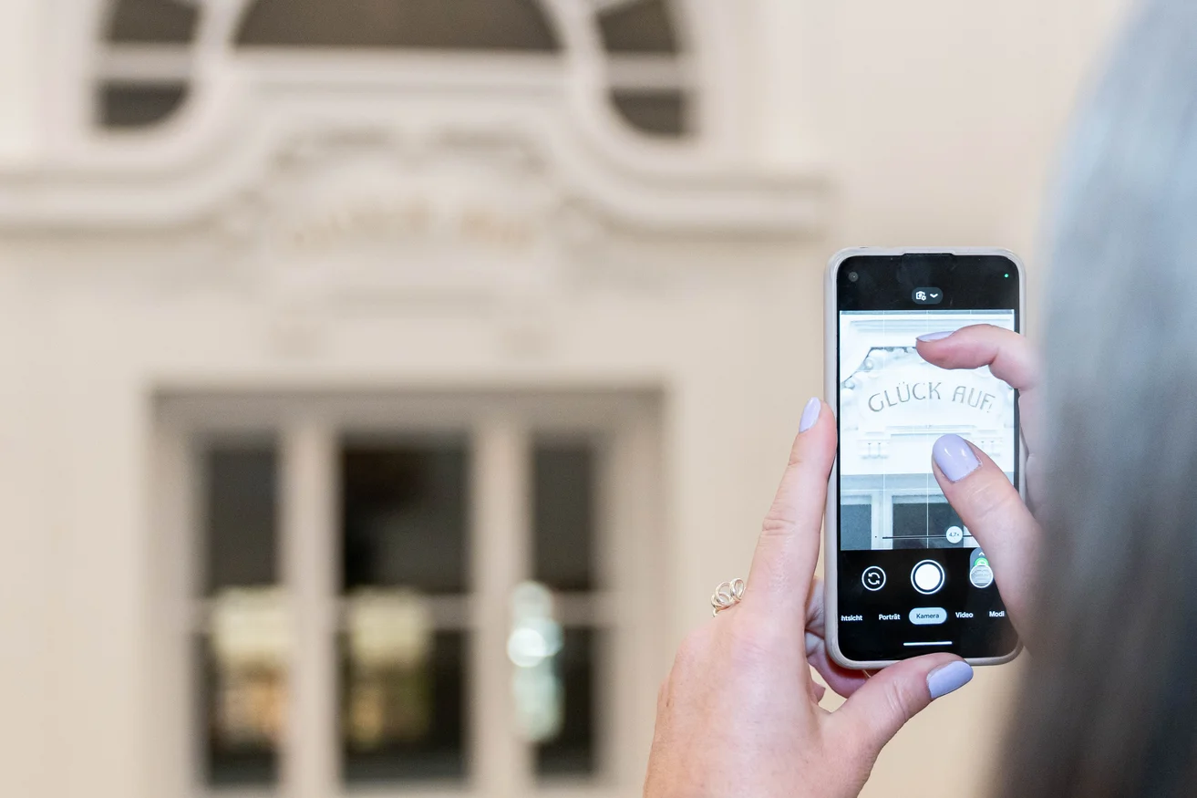 Eine Person fotografiert mit ihrem Smartphone den Leitspruch der Universität "Glück auf".