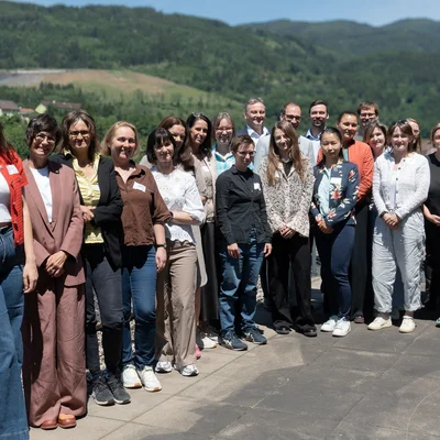 Gruppenfoto im Freien: Einige Personen stehen auf einer Terrasse mit Geländer und Solarpaneelen, dahinter grüne Hügel. Sonniger Tag.