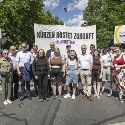 Eine Gruppe von Demonstrierenden steht auf einer Straße. Hinter ihnen hängt ein großes Banner mit der Aufschrift „Kürzen kostet Zukunft – #unisretten“. Rechts wird ein Schild „Mehr Bildung, mehr Wissen“ hochgehalten. Viele weitere Teilnehmende und Bäume im Hintergrund, sonniges Wetter.