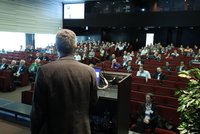 Redner am Podium spricht zu einem aufmerksam zuhörenden Publikum in einem Hörsaal.