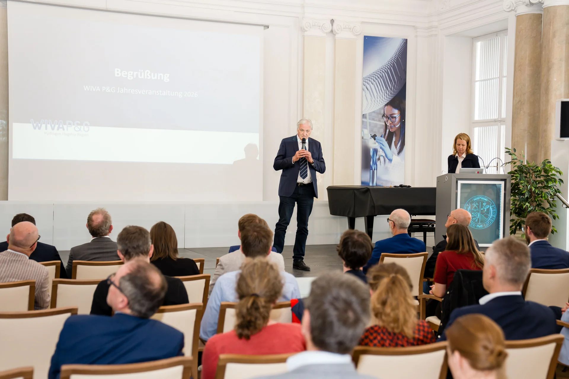 Konferenzszene in einem hellen Saal: Publikum sitzt in Reihen, vorne spricht eine Person mit Mikrofon vor einer Leinwand („Begrüßung – WIVA P&G Jahresveranstaltung 2026“). Rechts steht eine weitere Person am Rednerpult; im Hintergrund ein Flügel und Technik‑Banner.