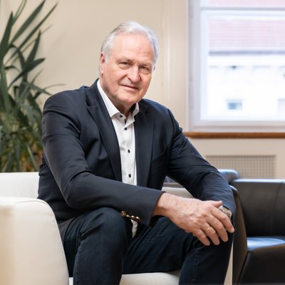 Rektor Peter Moser sitzt auf einem weißen Stuhl. Er trägt einen dunklen Anzug.