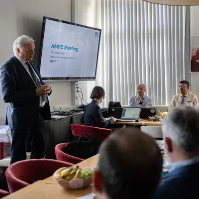 Konferenzraum mit langen Tischen: Teilnehmende sitzen rundum, einige mit Laptops; links präsentiert eine Person vor einem Bildschirm mit „AMRD Meeting … Agenda“. Große Pendelleuchten, vertikale Lamellenvorhänge.
