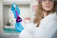 Frau in einem Laborkitteln mit einem Reagenzglas mit lila Flüssigkeit in der Hand.