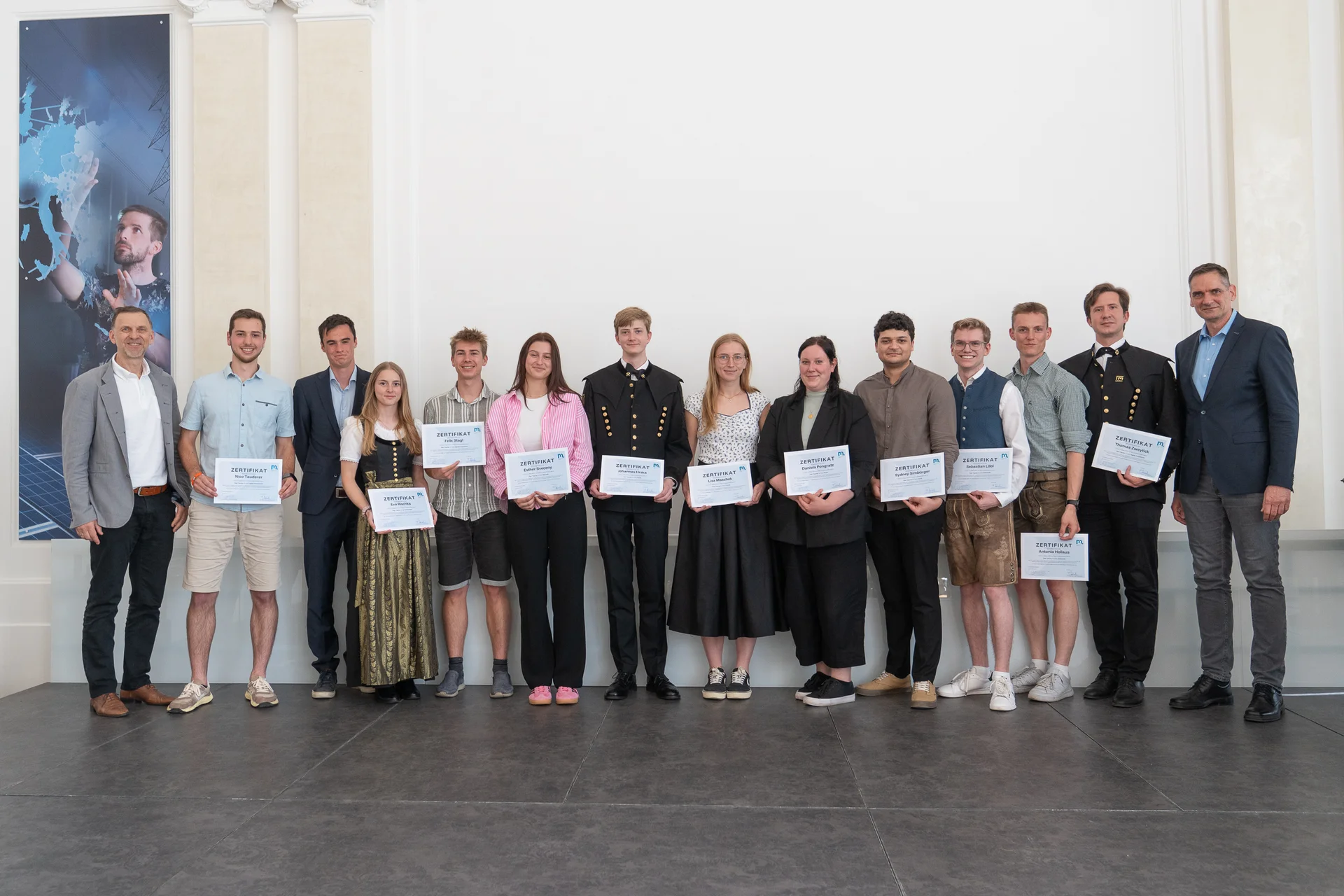 Gruppe von etwa zwölf Personen in einer hellen Halle, die für ein Foto posieren. Die meisten halten Zertifikate; Kleidung gemischt von Anzug bis casual, teils Uniform/Trachten. Veranstaltung wirkt wie eine Preis- oder Abschlussverleihung.