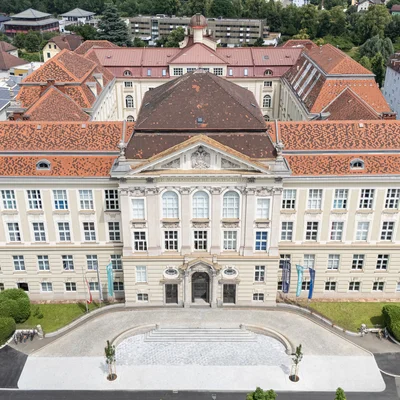 Luftaufnahme einer historischen Universitätsfassade mit roten Ziegeldächern; symmetrisches Hauptgebäude mit Portal, Flaggen am Vorplatz und umliegender Stadtbebauung.