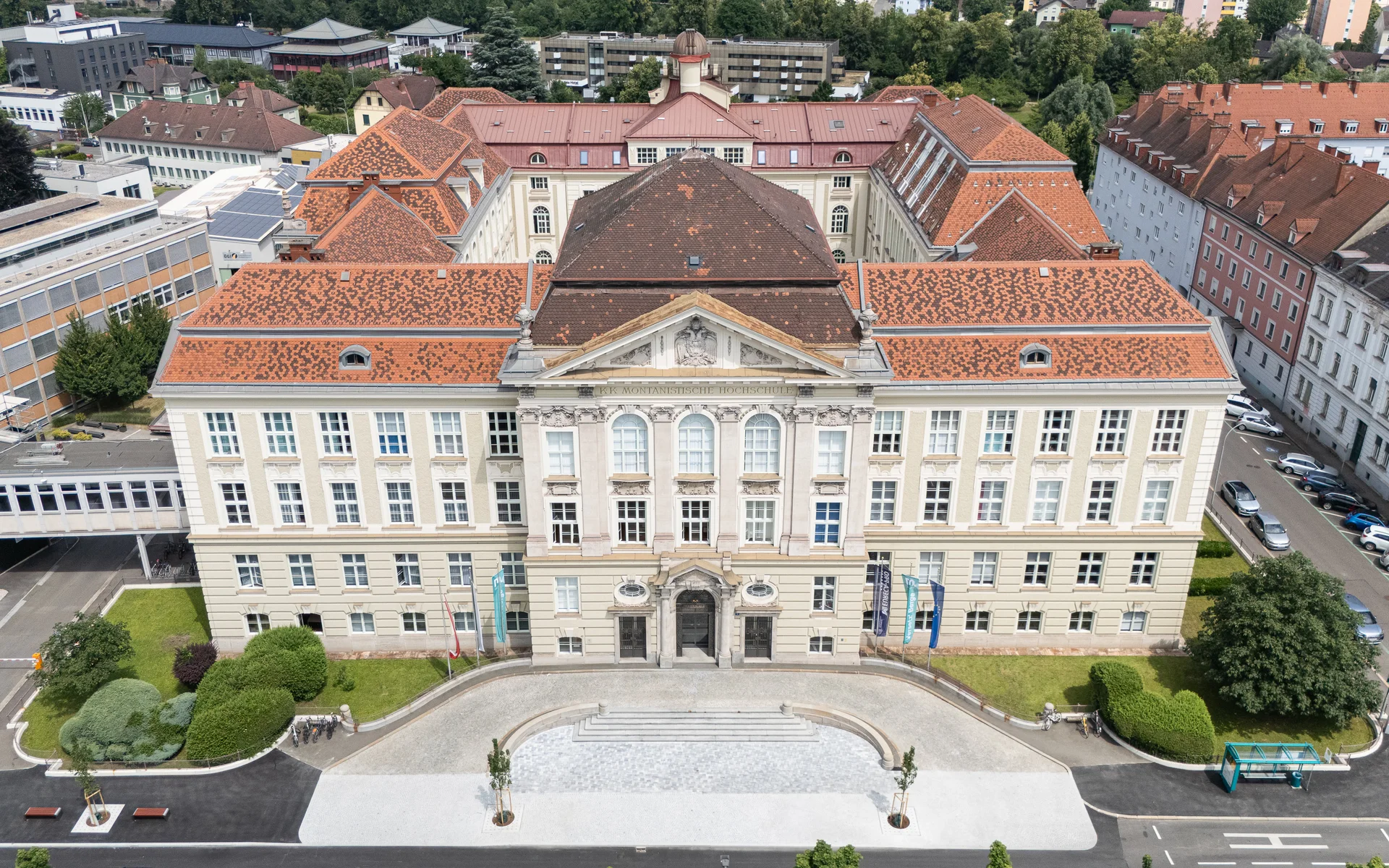 Luftaufnahme einer historischen Universitätsfassade mit roten Ziegeldächern; symmetrisches Hauptgebäude mit Portal, Flaggen am Vorplatz und umliegender Stadtbebauung.