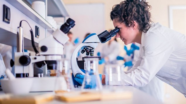 A woman in a white coat is in a laboratory looking through a microscope.
