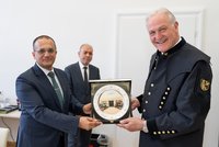 Zwei Männer überreichen lächelnd eine Ehrenplatte in Box; ein dritter im Hintergrund, formeller Rahmen.