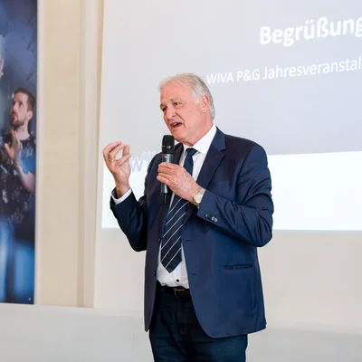 Eine Person im blauen Anzug spricht mit Mikrofon vor einer Leinwand mit der Folie „Begrüßung – WIVA P&G Jahresveranstaltung 2026“.