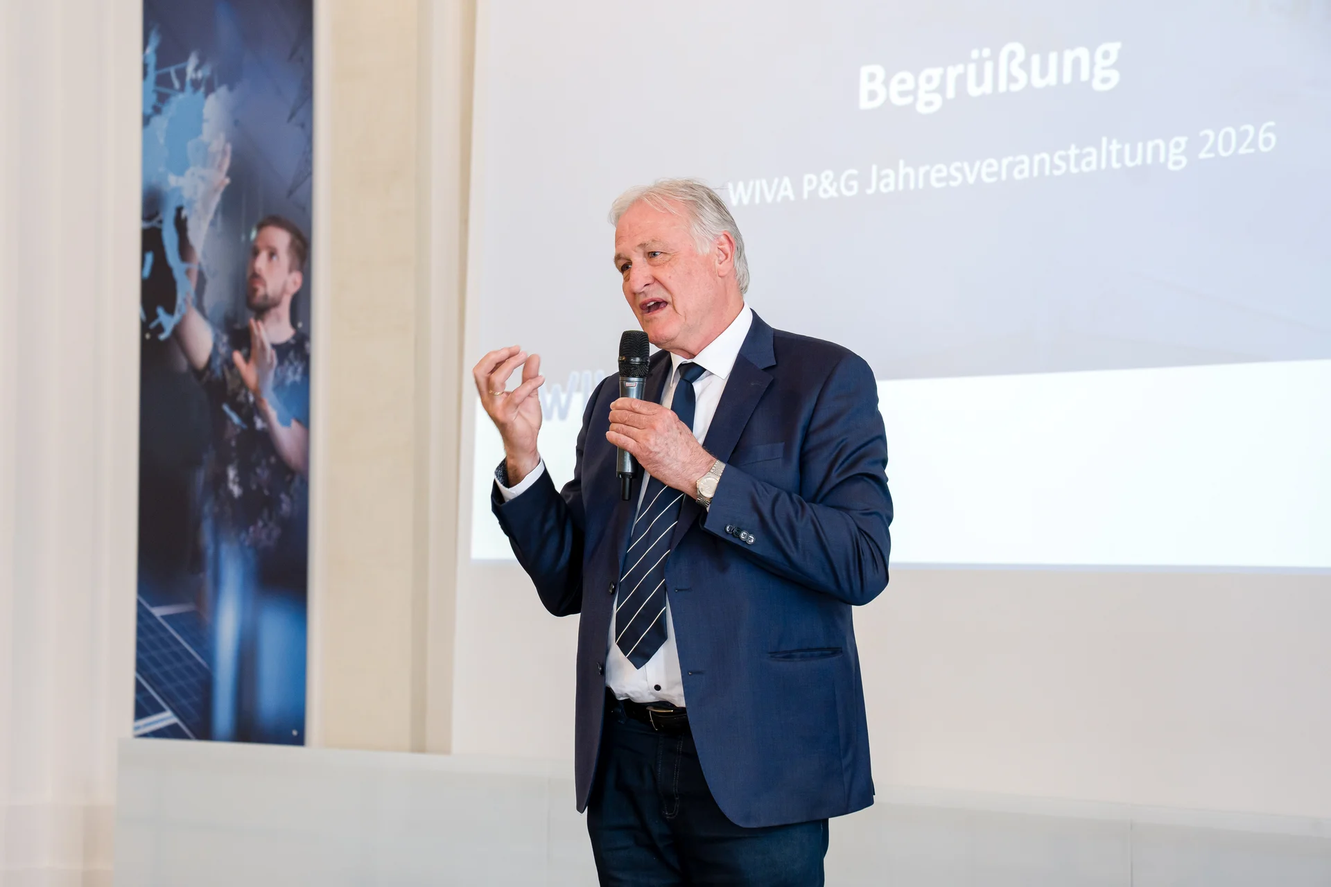Eine Person im blauen Anzug spricht mit Mikrofon vor einer Leinwand mit der Folie „Begrüßung – WIVA P&G Jahresveranstaltung 2026“.