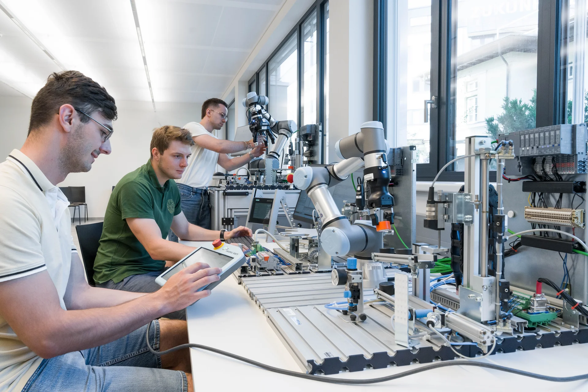 Mehrere Personen arbeiten in einem Labor an modularen Automatisierungsanlagen: kollaborative Roboterarme, Fördermodule und Sensorik auf Aluprofilen; eine Person bedient ein Tablet, andere justieren Geräte an Laptops nahe Fensterfront.