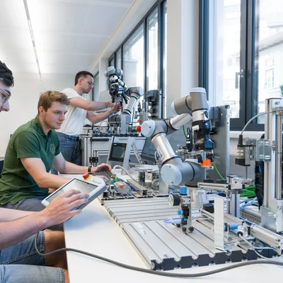Drei Personen arbeiten an einem Laborplatz mit kollaborativen Roboterarmen; eine steuert per Tablet, andere justieren Module und Sensoren.