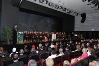 Ein großer Saal bei einer akademischen Feier: Publikum sitzt in Reihen, auf der Bühne eine Gruppe in festlicher Amtstracht, ein Redner am Pult, Beleuchtung von oben; Willkommensprojektion im Hintergrund.