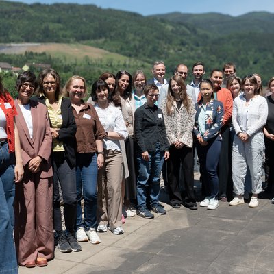 Gruppenfoto im Freien: Einige Personen stehen auf einer Terrasse mit Geländer und Solarpaneelen, dahinter grüne Hügel. Sonniger Tag.