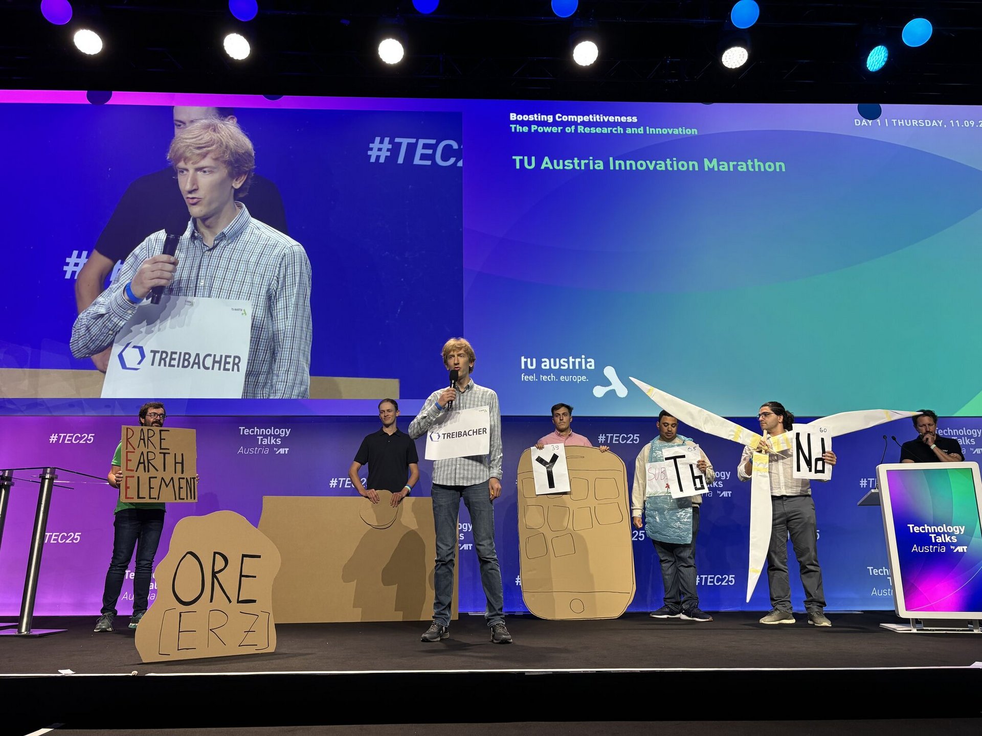 Präsentation der Ideen auf der Bühne: auf dem großen Screen direkt auf der Bühne, sieht man einen jungen Mann mit Mikro und einem umgehängten Schild mit "Treibacher". Dieser Mann steht auch mittig auf der Bühne. Im Hintergrund stehen 6 Personen mit Schildern. Eine Person hat ein selbst gebasteltes Windrad in der Hand.
