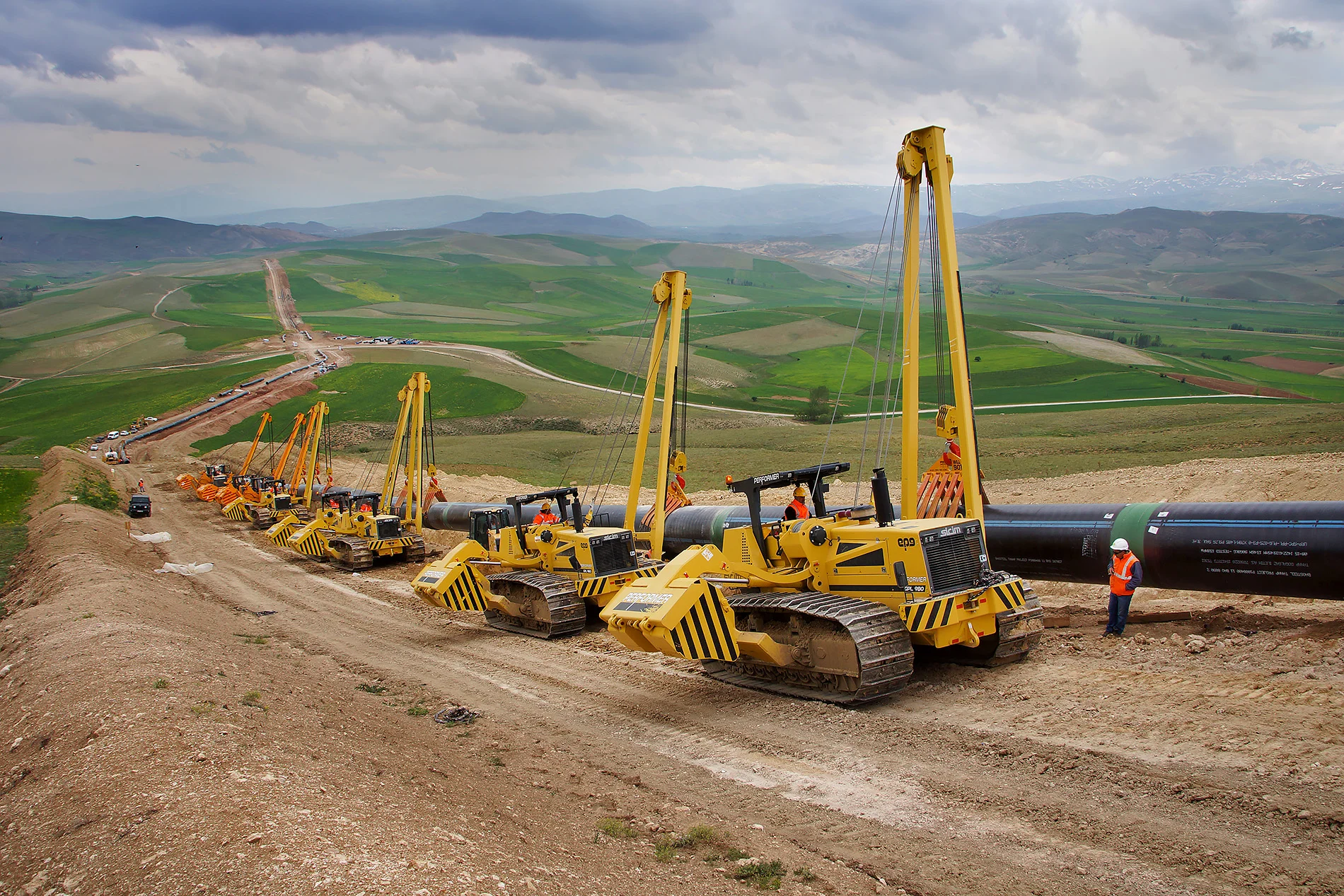Mehrere gelbe Rohrverleger heben und positionieren große Stahlrohre auf einer Hügelbaustelle; Arbeiter stehen neben der Pipeline.