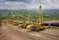 Mehrere gelbe Rohrverleger heben und positionieren große Stahlrohre auf einer Hügelbaustelle; Arbeiter stehen neben der Pipeline.