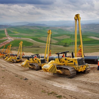 Mehrere gelbe Rohrverleger heben und positionieren große Stahlrohre auf einer Hügelbaustelle; Arbeiter stehen neben der Pipeline.