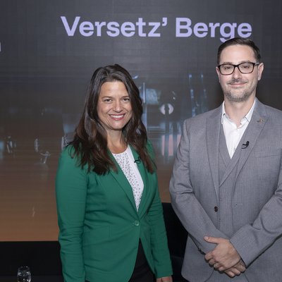 Zwei Personen in Business‑Kleidung stehen vor einer Bühnen‑/Event‑Leinwand mit dem Schriftzug „Versetz’ Berge“ und dem Logo der Montanuniversität Leoben. Links eine Person mit grünem Blazer, rechts eine Person im grauen Anzug.