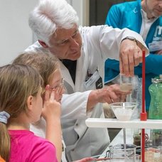 Forscher im Laborkittel zeigt Besuchern einen chemischen Versuch.